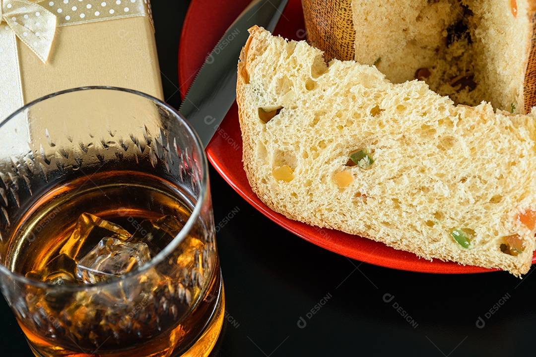 Closeup de uma fatia de panetone ao lado de uma faca, um copo com uísque e gelo e uma caixa de presente.