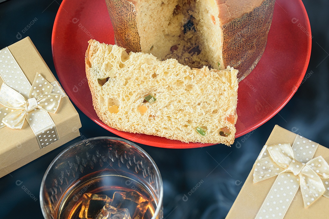 Close up da fatia de panetone sob a fumaça do cigarro, ao lado de um copo com uísque e gelo e caixas de presente.