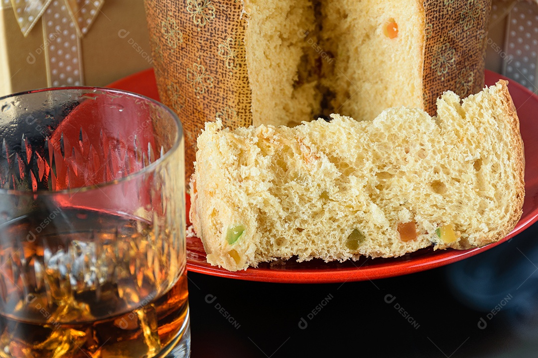 Fatia de panetone sob fumaça de cigarro, ao lado de um copo de uísque e gelo e caixa de presente
