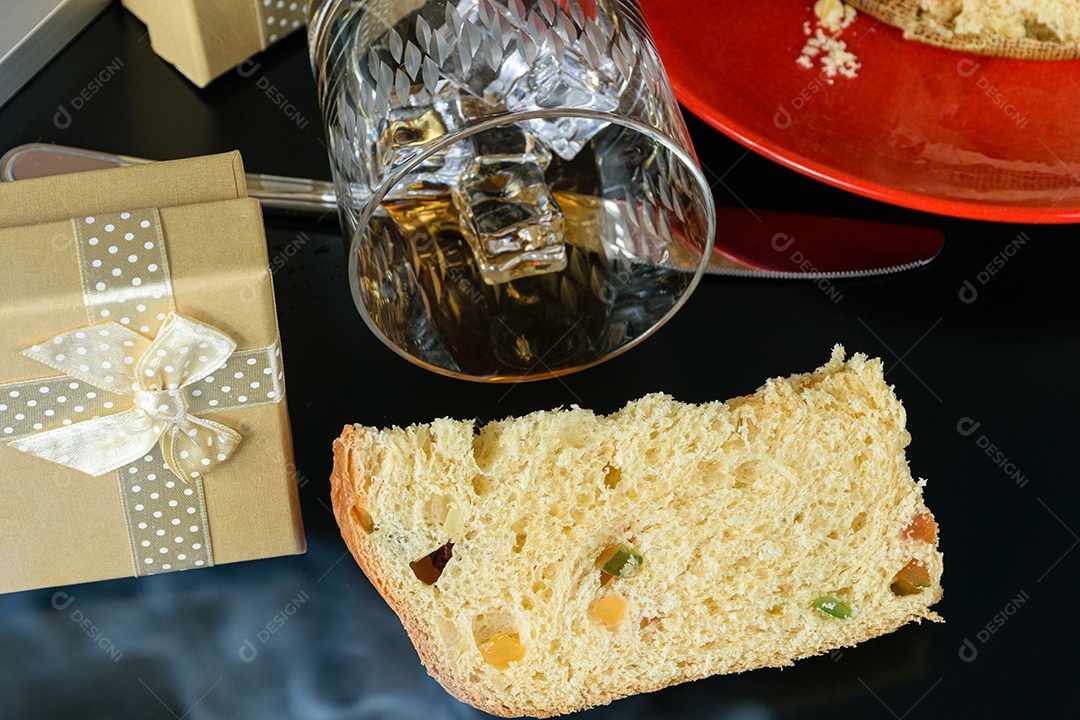 Fatia de panetone sob fumaça de cigarro, ao lado de uma caixa de presente e um copo de uísque