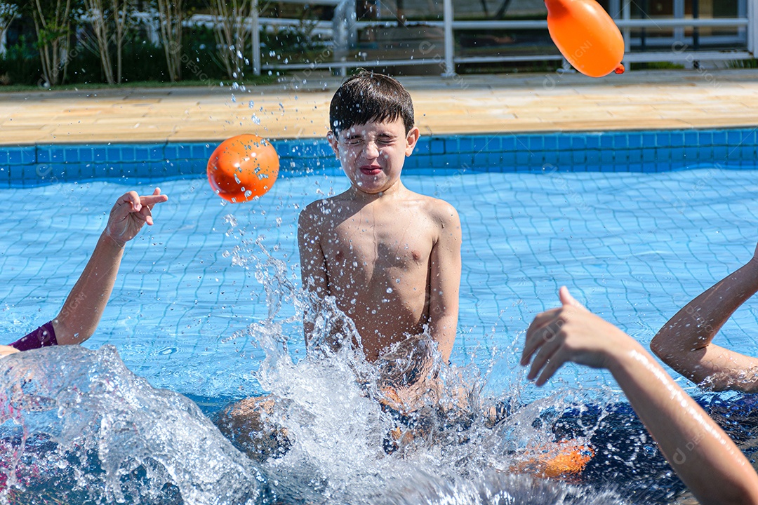 Criança sentada na beira da piscina, fazendo careta e quase sendo atingida por várias bombas de água.