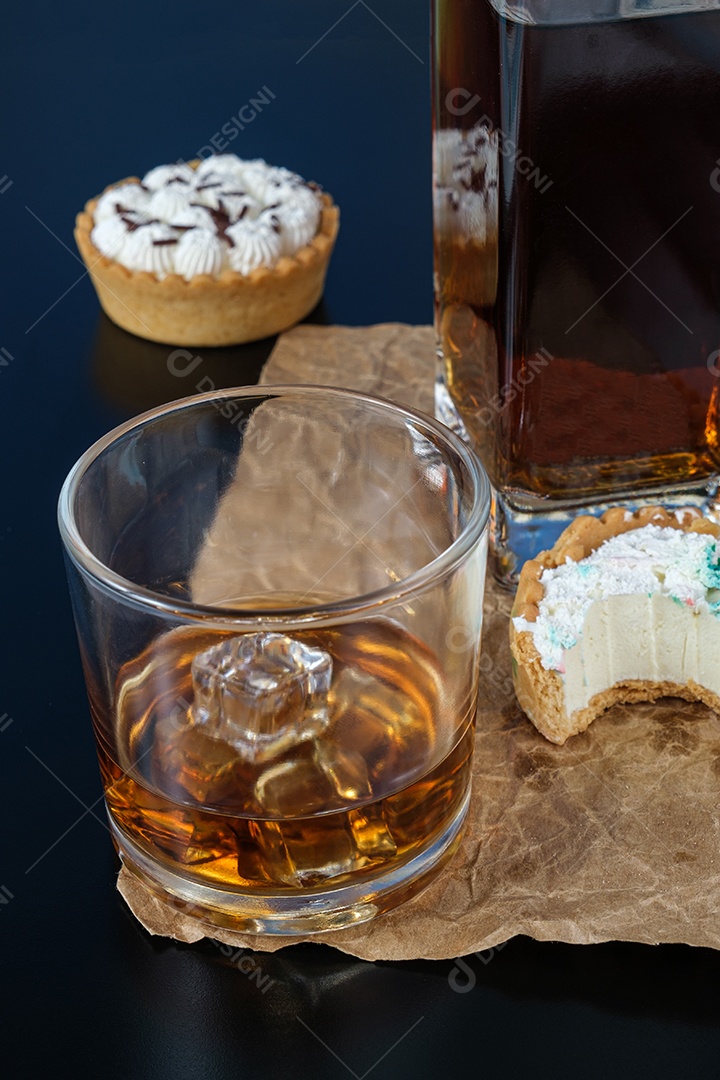 Copo de uísque com gelo, ao lado de uma mini torta, com uma mordida