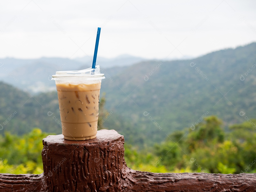 Um copo de café gelado é colocado em cima do muro em um cenário de montanhas e céu.