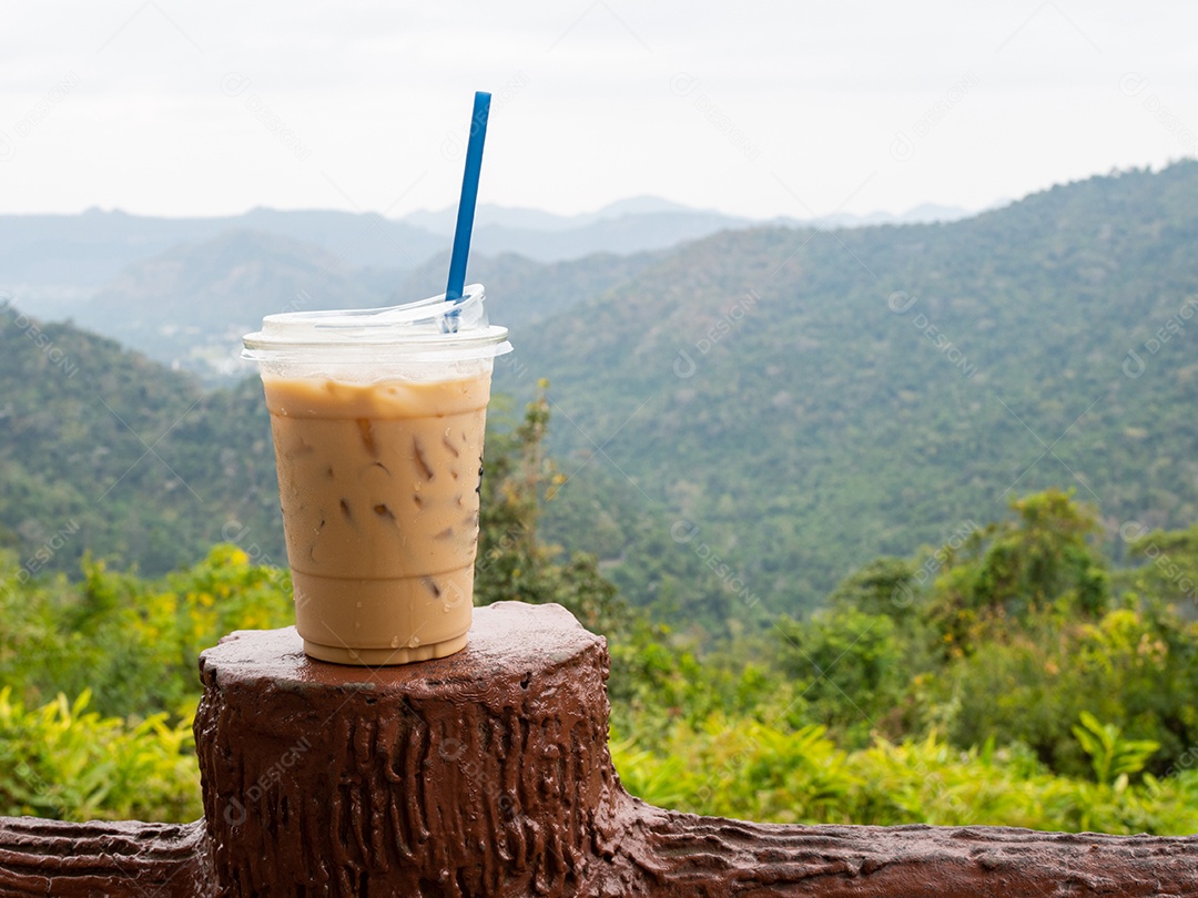 Um copo de café gelado é colocado em cima do muro em um cenário de montanhas e céu.