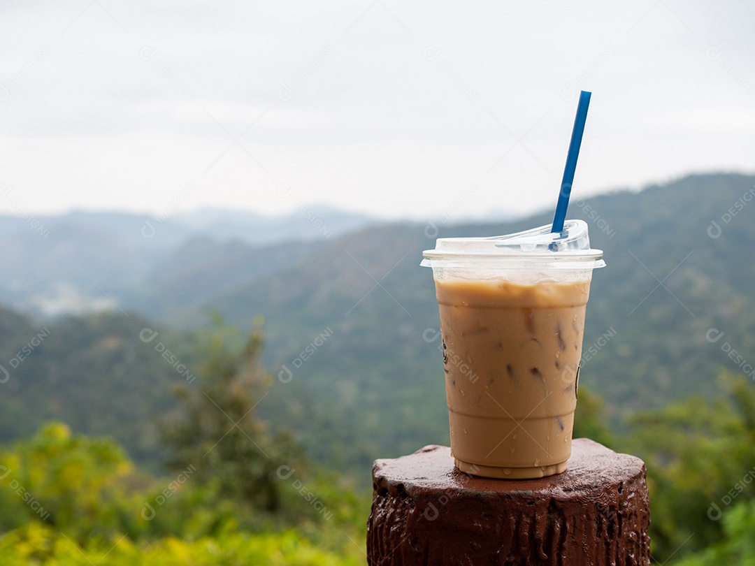 Um copo de café gelado é colocado em cima do muro em um cenário de montanhas e céu.