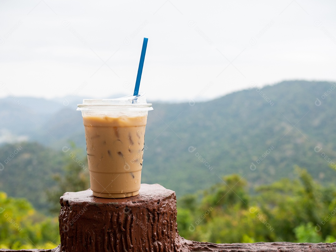 Um copo de café gelado é colocado em cima do muro em um cenário de montanhas e céu.