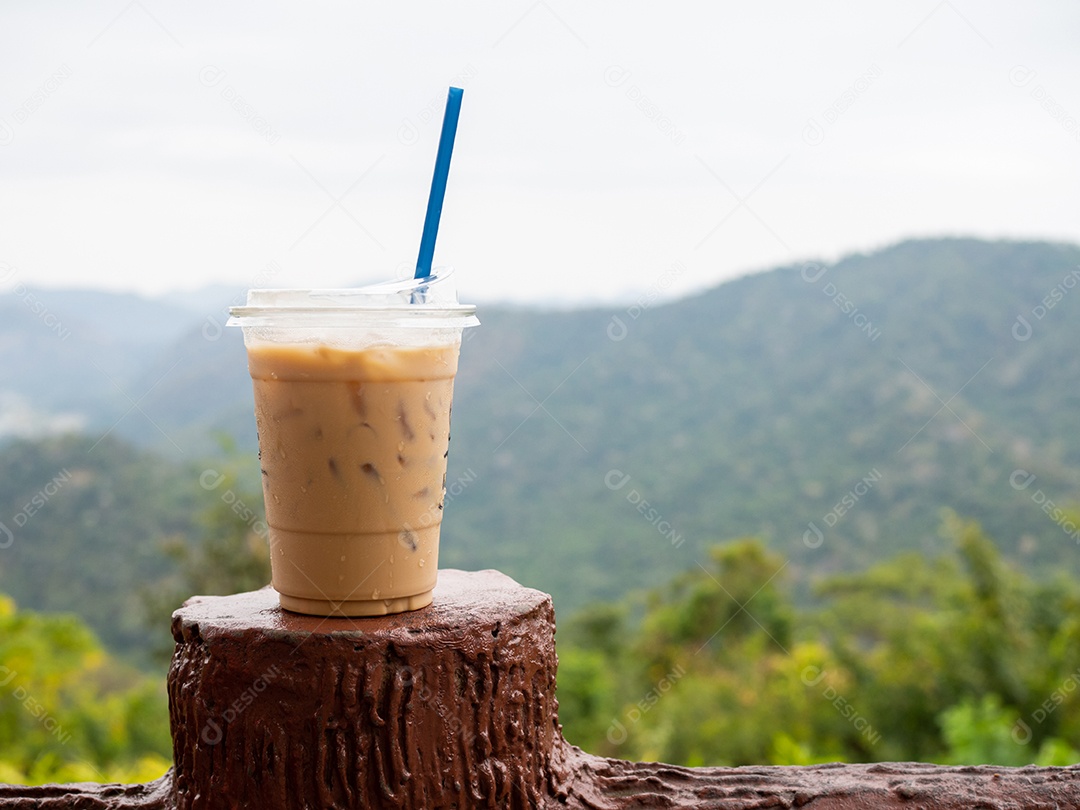 Um copo de café gelado é colocado em cima do muro em um cenário de montanhas e céu.