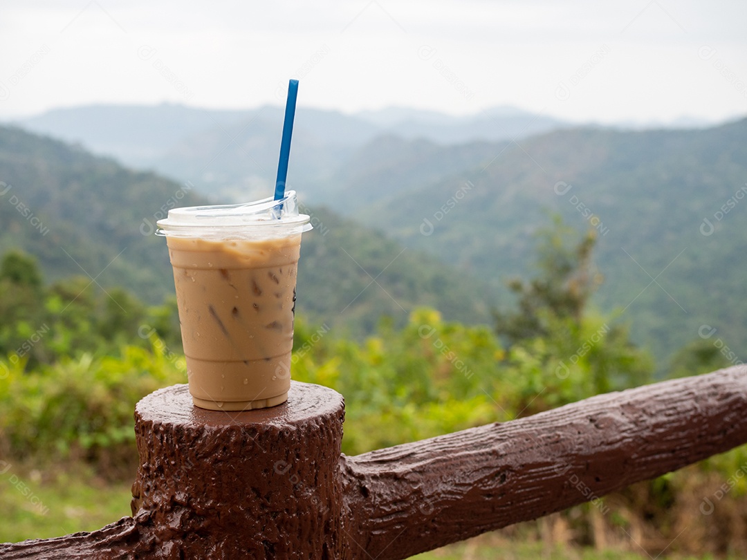 Um copo de café gelado é colocado em cima do muro em um cenário de montanhas e céu.