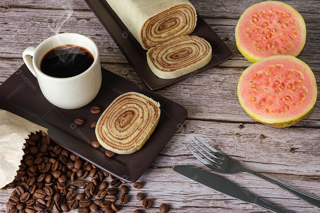 Fatia de bolo de rolo rodeada de talheres, goiabas, xícara e café