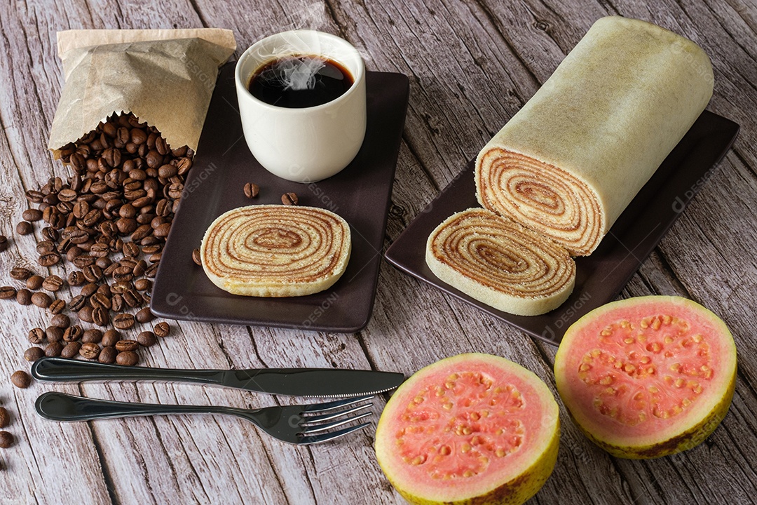 Fatia de bolo de rolo rodeada de talheres, goiabas, xícara e café