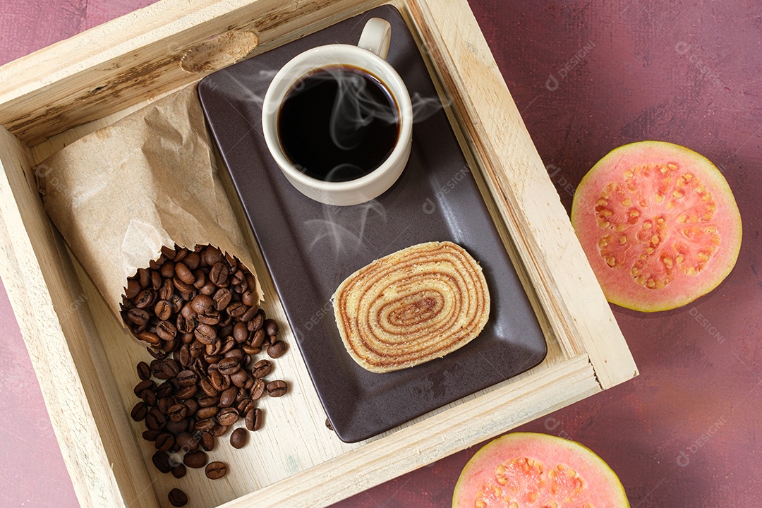 Fatia de bolo de rolo rodeada de goiabada, xícara e grãos de café.
