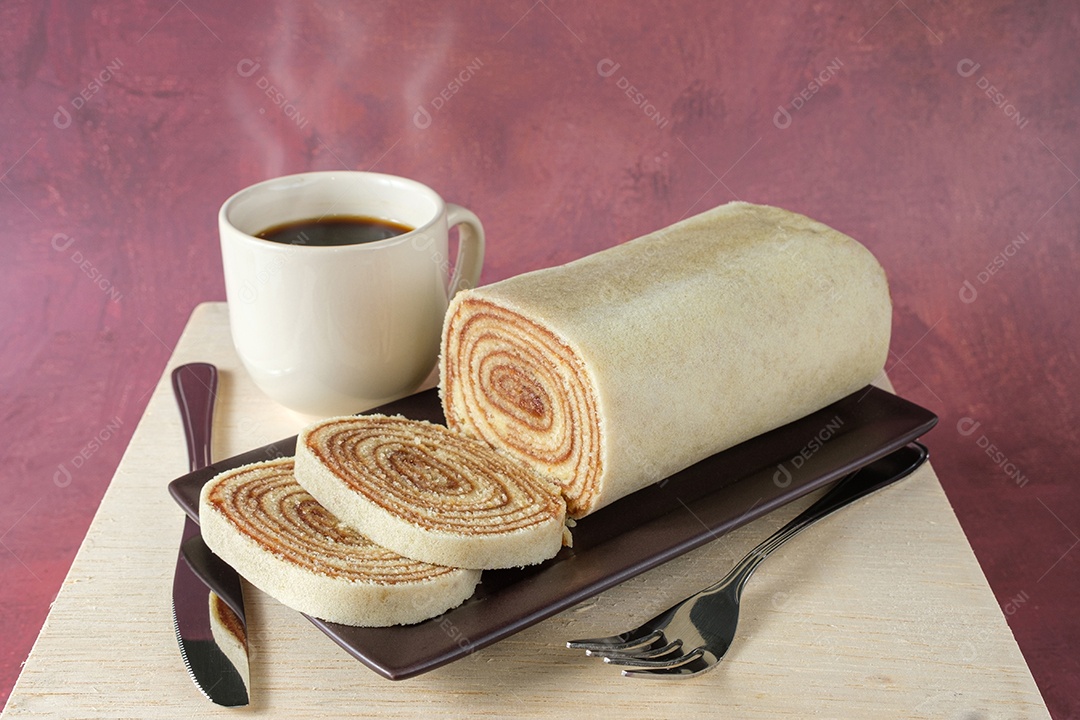 Bolo de rolo fatiado em um prato marrom, ao lado de talheres e uma xícara de café
