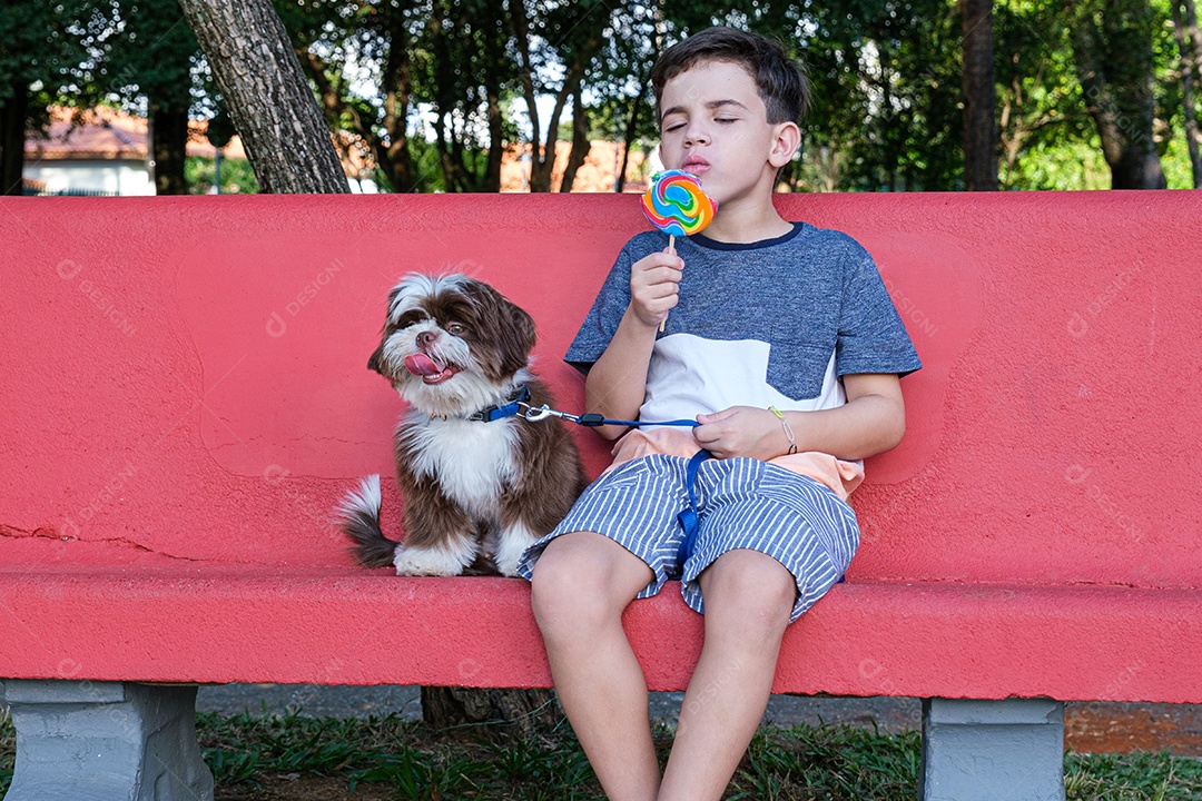 Menino de 8 anos sentado em um banco vermelho com um pirulito na mão