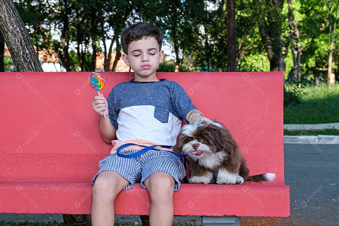 Menino de 8 anos sentado em um banco vermelho com um pirulito na mão