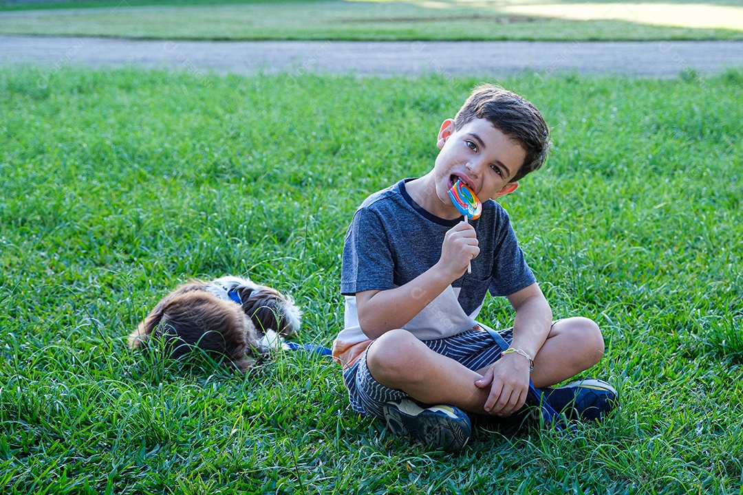 Menino de 8 anos sentado no gramado ao lado de seu animal de estimação e mordendo um pirulito