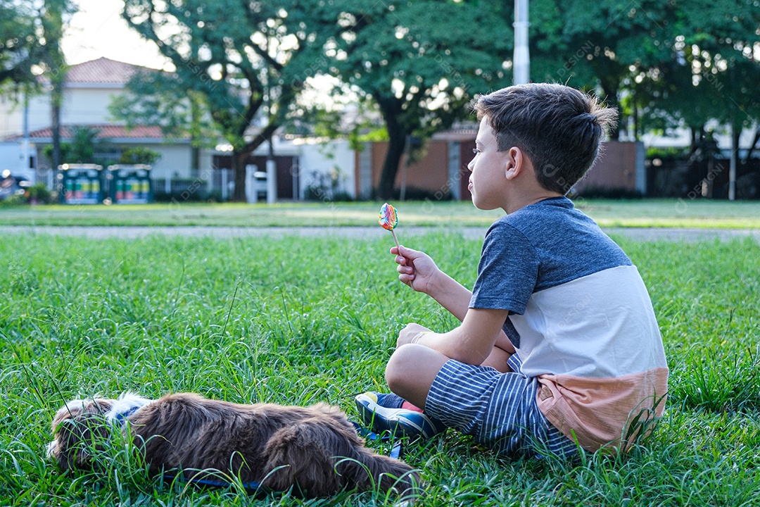 Menino de 8 anos olhando para a frente, sentado no gramado ao lado de seu cachorro