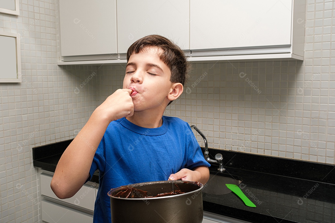 Criança de 8 anos na cozinha segurando uma panela, olhando para cima e lambendo o dedo com brigadeiro.