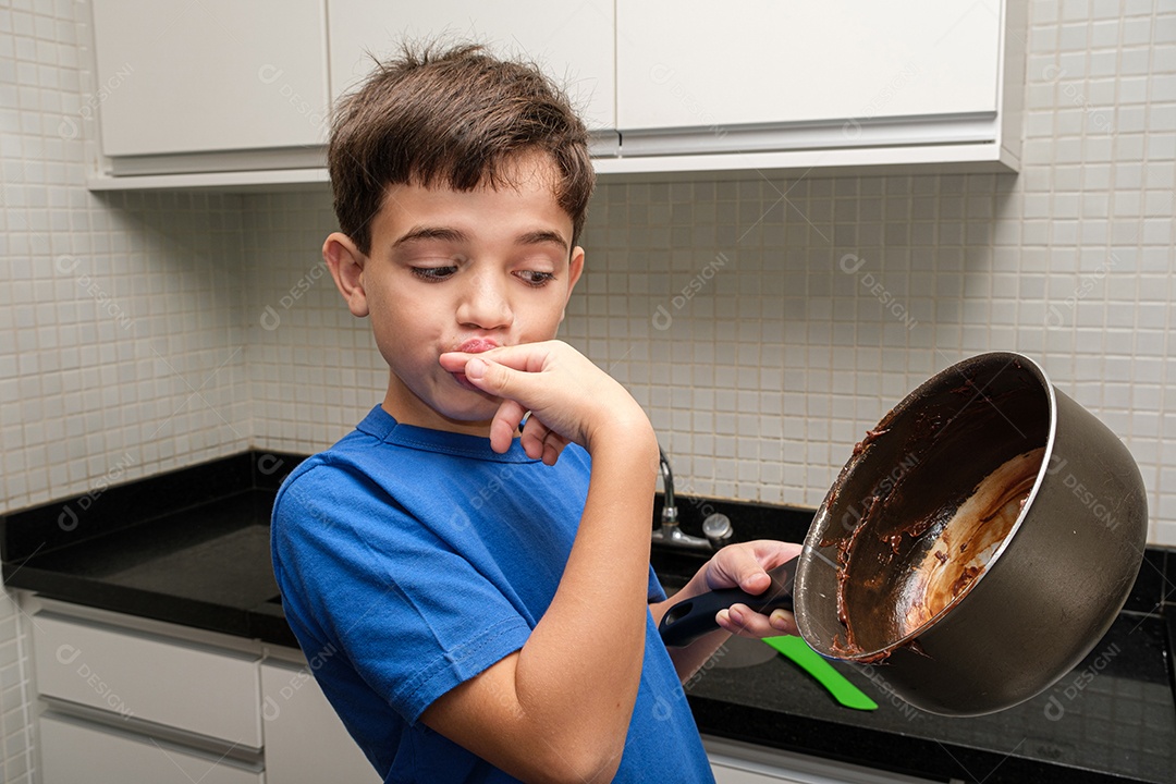 Criança de 8 anos na cozinha lambendo o dedo e olhando a panela com brigadeiro.