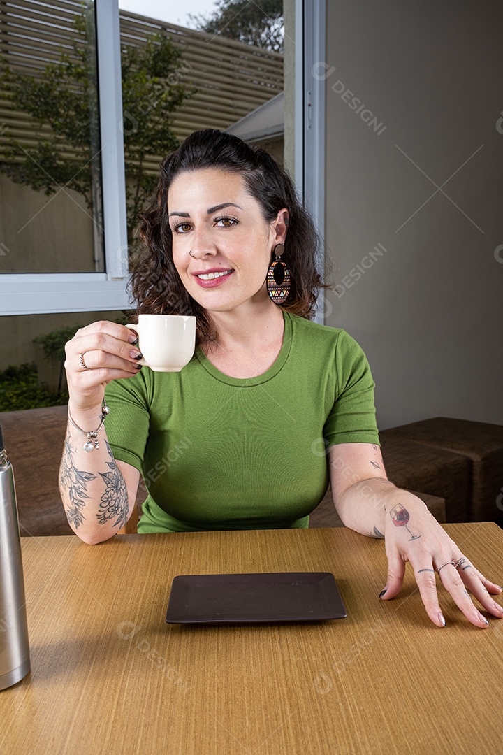 Mulher brasileira sorridente e tatuada sentada e segurando uma xícara