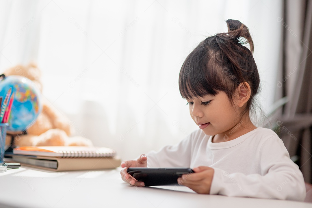 Menina asiática usando um telefone de perto, distraída de estudar, sentada em uma mesa com notebooks, uma criança bonita se divertindo com um smartphone, assistindo ao webinar, educação em casa