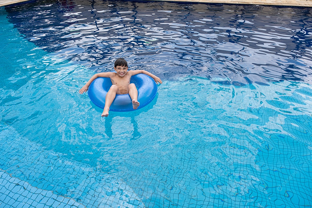 Brasileiro de 9 anos na piscina, fazendo careta, em uma boia em um dia ensolarado.