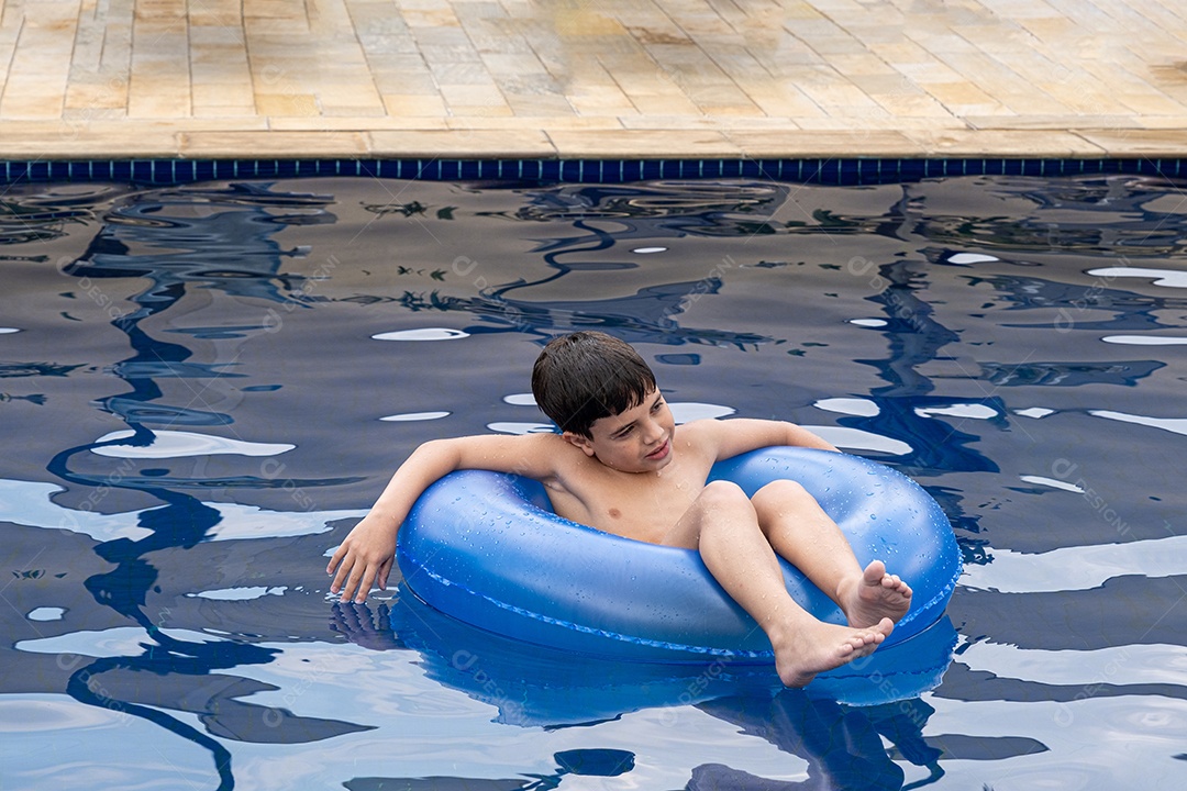 Brasileiro de 9 anos na piscina, fazendo careta, em uma boia em um dia ensolarado.