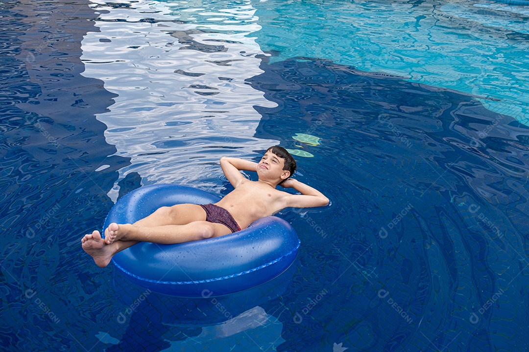 Brasileiro de 9 anos na piscina, fazendo careta, em uma boia em um dia ensolarado.