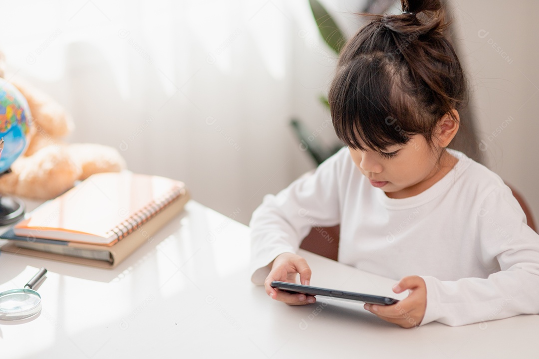 Menina asiática usando um telefone de perto, distraída de estudar, sentada em uma mesa com notebooks, uma criança bonita se divertindo com um smartphone, assistindo ao webinar, educação em casa