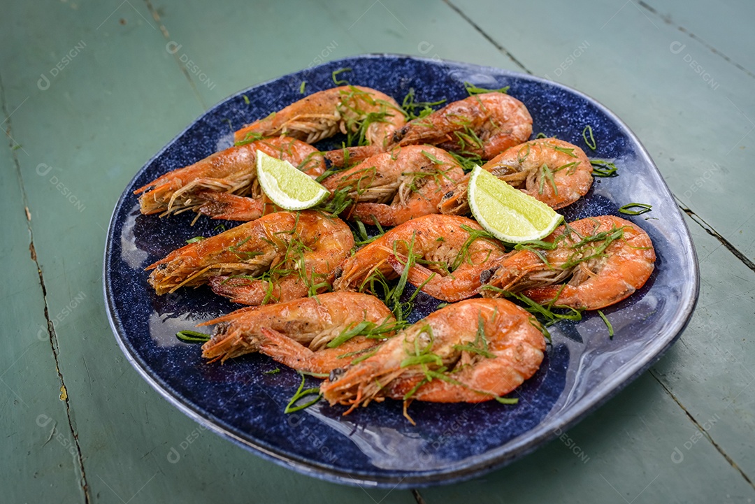 Camarão salteado com alho e azeite num prato azul escuro sobre uma mesa de madeira pintada de verde. Comida brasileira muito consumida no litoral.