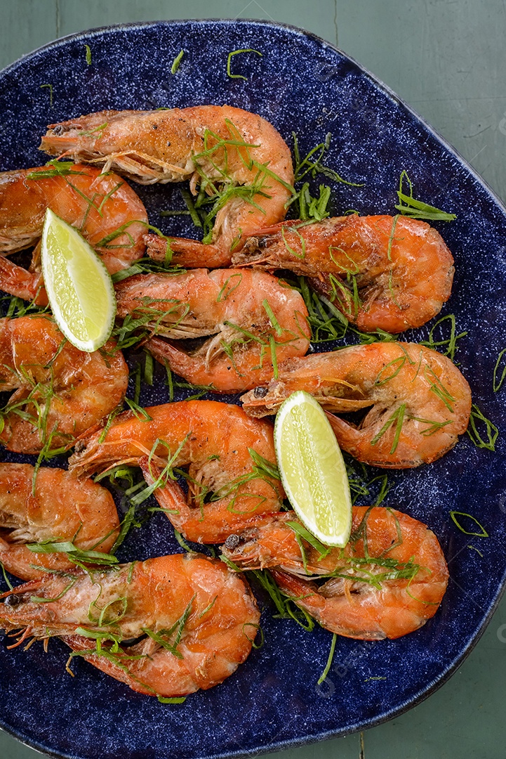 Camarão salteado com alho e azeite num prato azul escuro sobre uma mesa de madeira pintada de verde. Comida brasileira muito consumida no litoral.