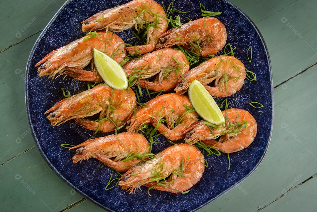 Camarão salteado com alho e azeite num prato azul escuro sobre uma mesa de madeira pintada de verde. Comida brasileira muito consumida no litoral.