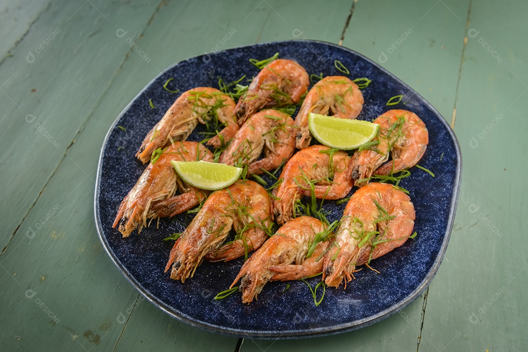 Camarão salteado com alho e azeite num prato azul escuro sobre uma mesa de madeira pintada de verde. Comida brasileira muito consumida no litoral.
