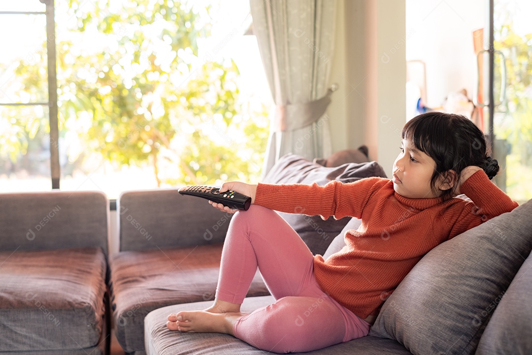 Menina asiática está assistindo TV e pressionando o controle remoto no sofá cinza na sala de estar perto da janela em casa, conceito de conforto na vida cotidiana