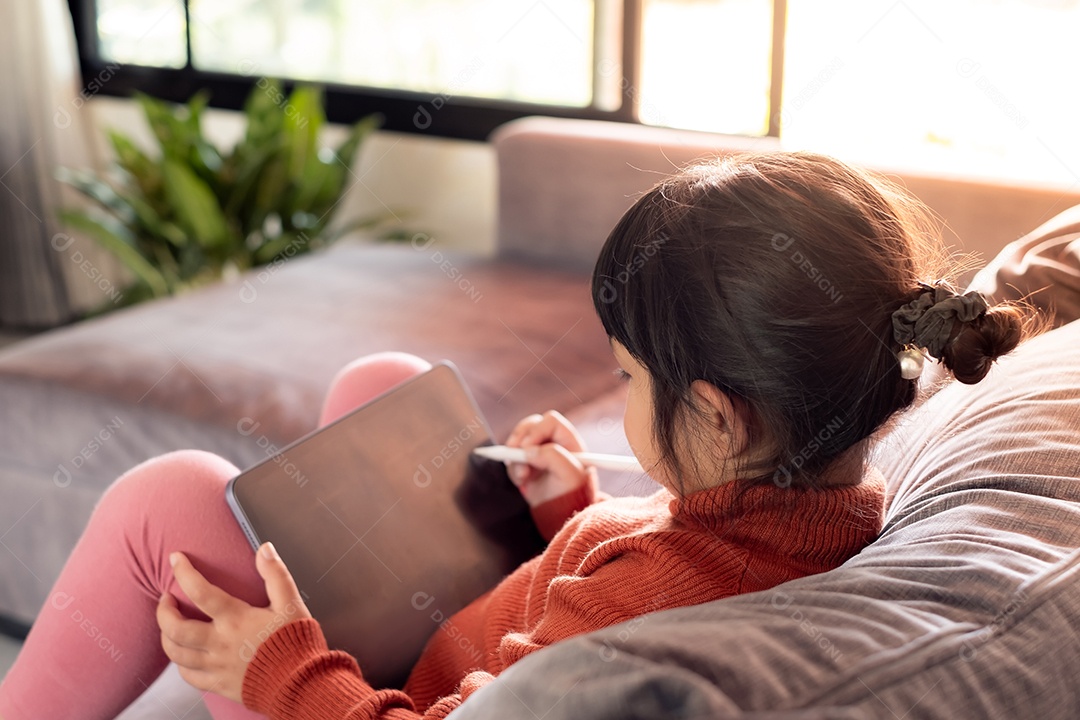 Retrato de uma linda menina asiática usando tablet para educação e configuração no sofá cinza perto da janela em casa, conceito de educação a distância em casa