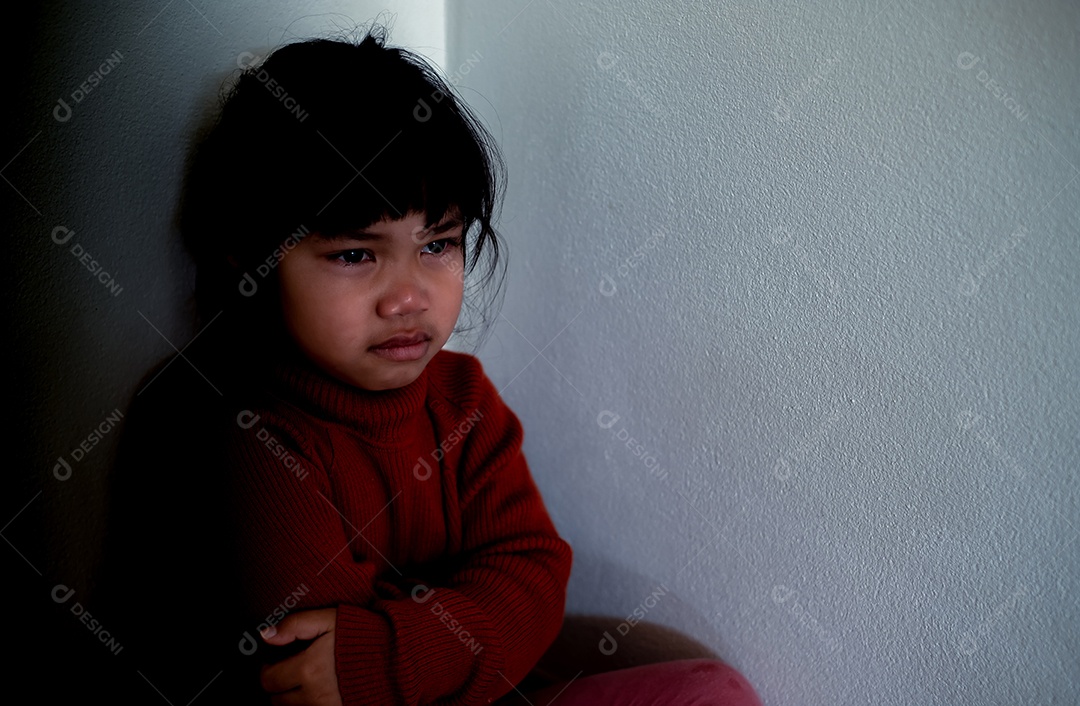 Menina asiática de 4 a 5 anos vestindo camisa vermelha está tristemente sentada ao lado de uma parede branca em uma sala mal iluminada, deprimida, solitária, estressada, chorando.