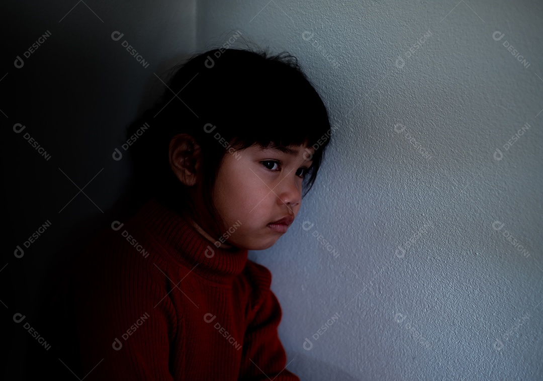 Menina asiática de 4 a 5 anos vestindo camisa vermelha está tristemente sentada ao lado de uma parede branca em uma sala mal iluminada, deprimida, solitária, estressada, chorando.