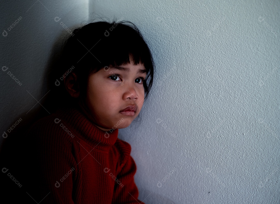 Menina asiática de 4 a 5 anos vestindo camisa vermelha está tristemente sentada ao lado de uma parede branca em uma sala mal iluminada, deprimida, solitária, estressada, chorando.