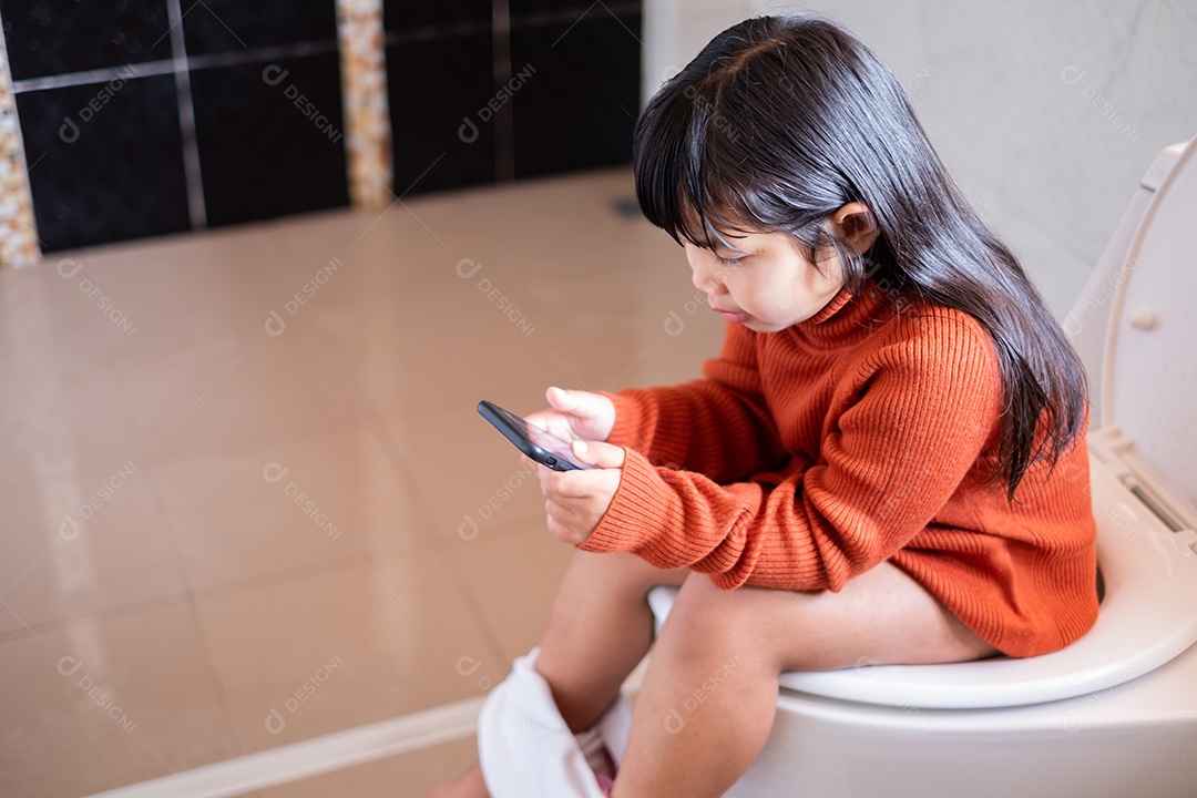 Linda garota asiática de pijama sentada no banquinho no vaso sanitário de manhã depois de acordar e olhar para o smartphone, problema infantil viciado em conceito de tecnologia