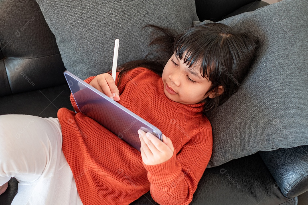 Retrato de uma linda menina asiática usando tablet para educação e configuração no sofá cinza perto da janela em casa, conceito de educação a distância em casa, vista superior