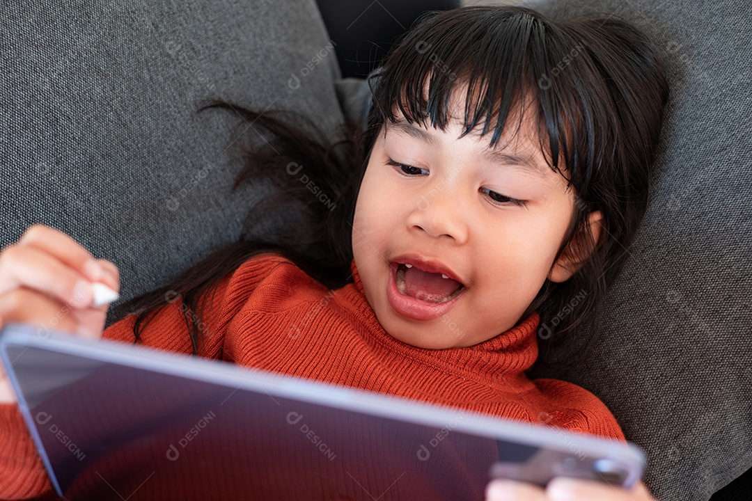 Retrato de uma linda menina asiática usando tablet para educação e configuração no sofá cinza perto da janela em casa, conceito de educação a distância em casa, vista superior