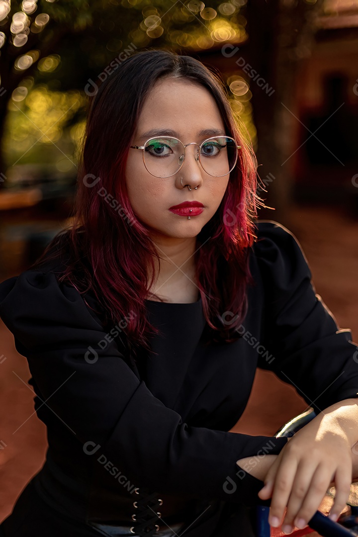 Jovem caucasiana de cabelos escuros com listras vermelhas em roupa de vestido preto com cinto espartilho e óculos posando para sessão de beleza feminina.