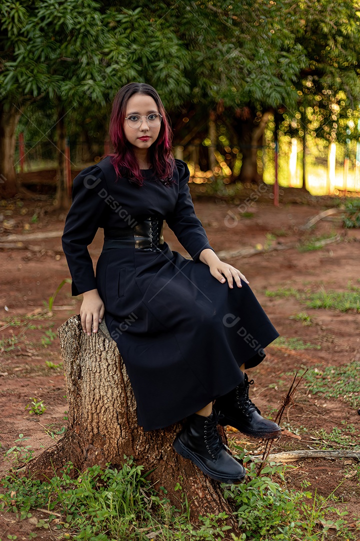 Jovem caucasiana de cabelos escuros com listras vermelhas em roupa de vestido preto com cinto espartilho e óculos posando para sessão de beleza feminina