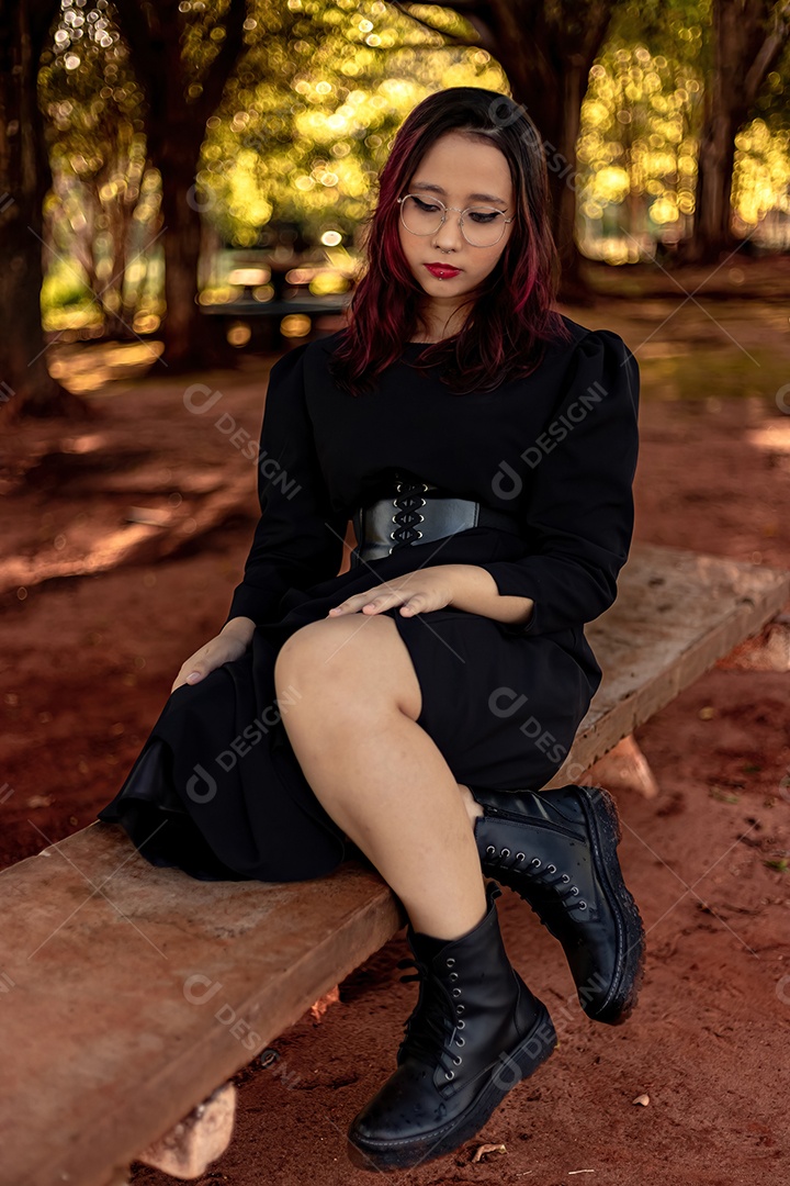 Jovem caucasiana de cabelos escuros com listras vermelhas em roupa de vestido preto com cinto espartilho e óculos posando para sessão de beleza feminina.