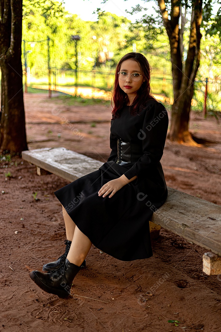 Jovem caucasiana de cabelos escuros com listras vermelhas em roupa de vestido preto com cinto espartilho e óculos posando para sessão de beleza feminina.