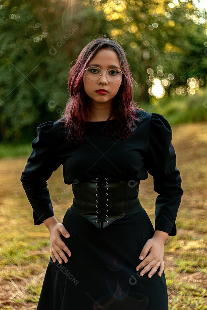 Jovem caucasiana de cabelos escuros com listras vermelhas em roupa de vestido preto com cinto espartilho e óculos posando para sessão de beleza feminina