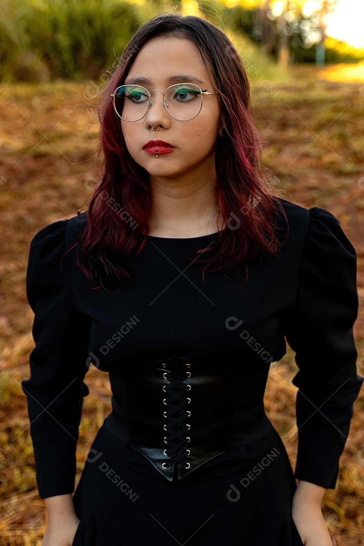 Jovem caucasiana de cabelos escuros com listras vermelhas em roupa de vestido preto com cinto espartilho e óculos posando para sessão de beleza feminina