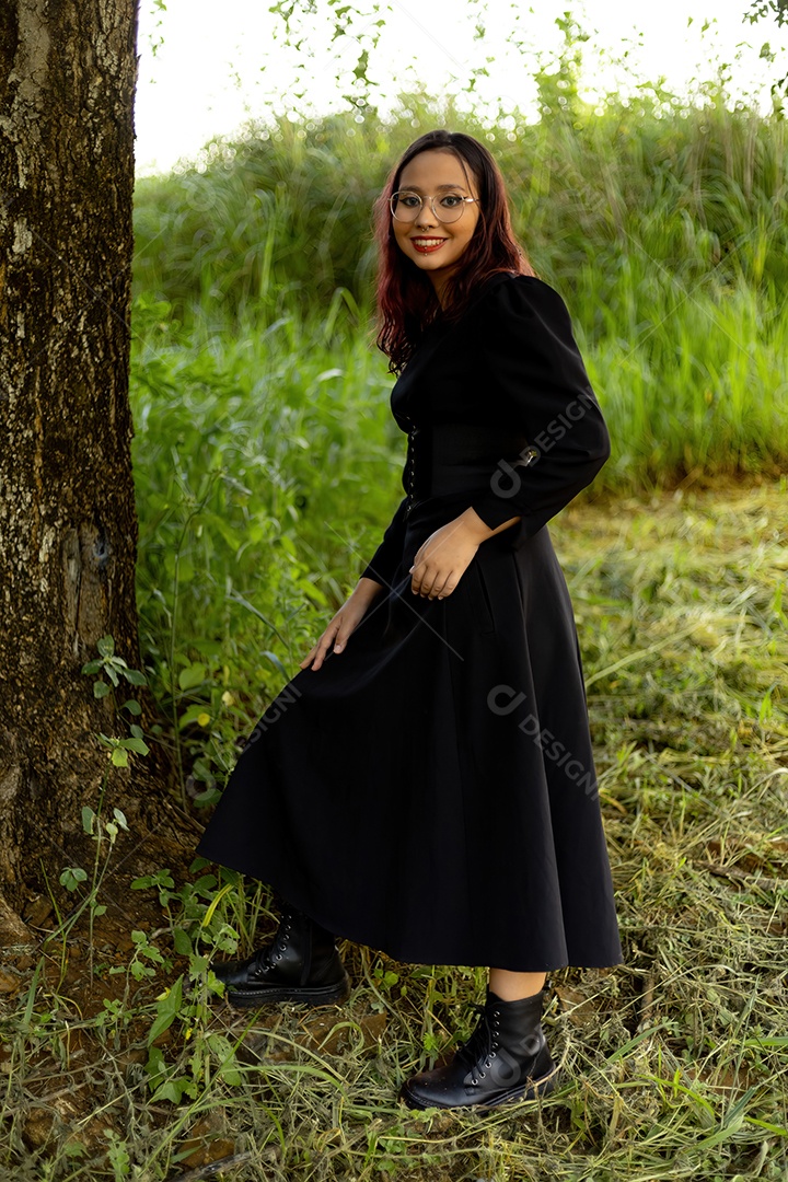 Jovem caucasiana de cabelos escuros com listras vermelhas em roupa de vestido preto com cinto espartilho e óculos posando para sessão de beleza feminina