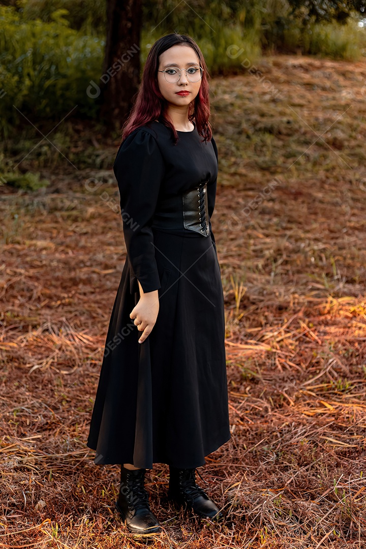 Jovem caucasiana de cabelos escuros com listras vermelhas em roupa de vestido preto com cinto espartilho e óculos posando para sessão de beleza feminina