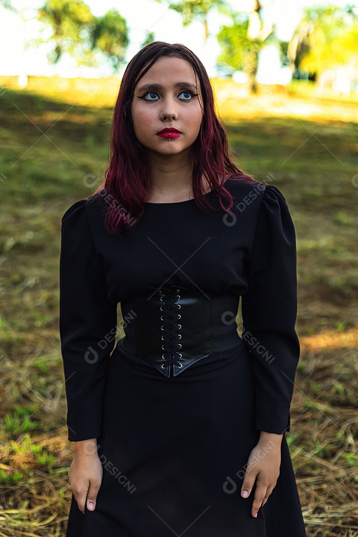 Jovem caucasiana de cabelos escuros com listras vermelhas em roupa de vestido preto com cinto espartilho e óculos posando para sessão de beleza feminina