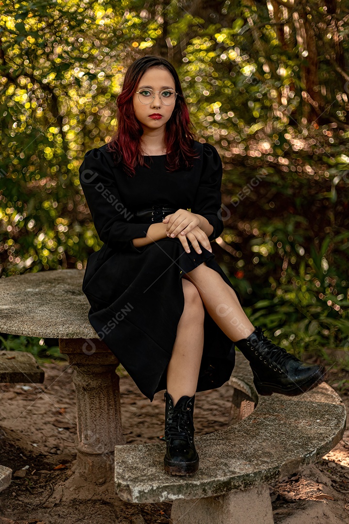 Jovem caucasiana de cabelos escuros com listras vermelhas em roupa de vestido preto com cinto espartilho e óculos posando para sessão de beleza feminina